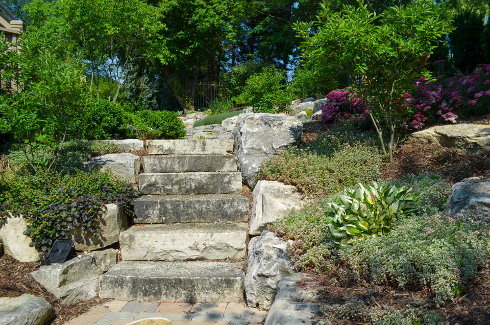 Stone stair run through terraced planting with limestone walls, boulders, shrubs, ornamental grasses, and wooded backdrop.
